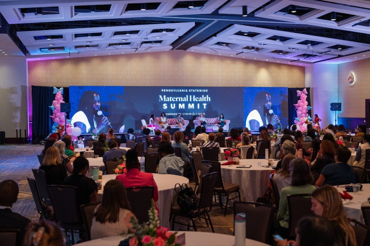 Large room of attendees sitting at tables and looking towards the stage.